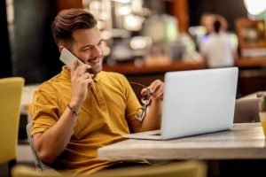 Un hombre atrae clientes mientras habla por teléfono sentado en una mesa con una computadora portátil.