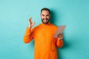 Un hombre con un suéter naranja está haciendo un signo de aprobación mientras sostiene una tableta, captando la atención de clientes potenciales para una campaña de anuncios de Facebook.